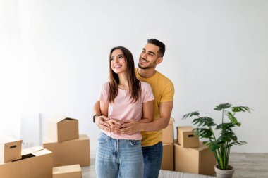 Genç çift gülümsüyor ve yeni evlerinde birbirlerini kucaklıyor, taşınan kutular ve yeşil bir bitkiyle çevrili. Paketleri açarken mutluluğu paylaşıyorlar..