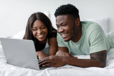 Bir erkek ve kadın, yatakta yatarken mutlu bir anı paylaşıyor, gülümsüyor ve dizüstü bilgisayarla meşgul oluyorlar. Oda aydınlık ve rahat, birlikte geçirdikleri zaman için rahat bir atmosfer yaratıyor..