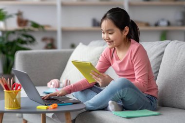 Neşeli bir kız dizüstü bilgisayarına odaklanır, açık gri bir koltukta dinlenirken sarı bir not alır. Odada bitkiler ve renkli okul malzemeleri var..