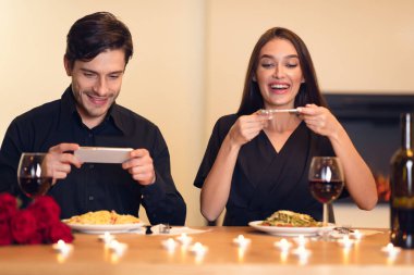 İki kişi, mumlarla süslenmiş bir masada oturmuş akşam yemeklerini belgelerken gülümsüyorlar. Makarna tabakları ve kırmızı şarap kadehleri mevcuttur, sıcak bir akşam olur..