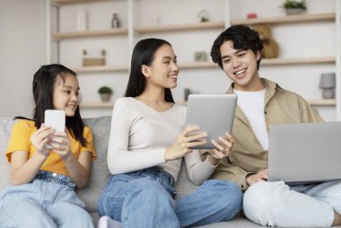 Üç kardeş bir kanepede rahatça oturur ve cihazlarıyla meşgul olurlar. Genç kız bir akıllı telefon kullanırken, erkek ve kız kardeşi bir tableti paylaşıyor. Mutlu ve odaklanmış görünüyorlar..