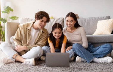 Genç bir kız ailesi ile yerde oturuyor, dizüstü bilgisayara odaklanmış. Gülümsüyorlar ve rahat oturma odalarında birlikte mutlu bir anı paylaşıyorlar..