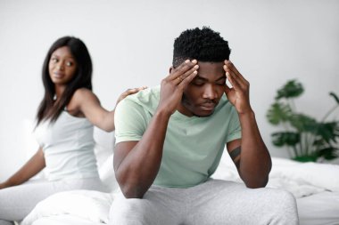 Bir kadın omzuna dokunurken, bir erkek acı dolu bir ifadeyle yatağın kenarına oturur. Sıcak, aydınlık bir odada duygusal bir çatışma yaşıyor gibi görünüyorlar..