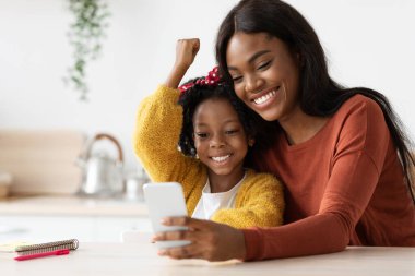 Neşeli bir anne ve kız, akıllı telefon ekranına bakıp gülümseyen ve kutlama yapan özel bir anı paylaşıyorlar. Parlak bir mutfak masasında oturuyorlar..