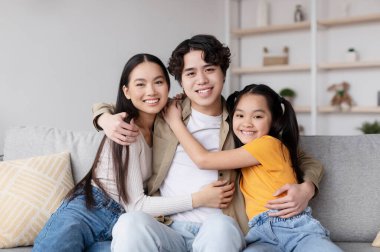 Genç bir aile oturma odasında keyifli bir anı paylaşıyor. Kanepede oturuyorlar, gülümsüyorlar ve birbirlerine sarılıyorlar. Hava sıcak ve davetkar..