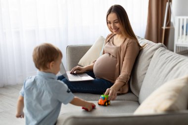 Hamile bir kadın, parlak bir oturma odasındaki koltukta oturuyor ve yakınlardaki oyuncak arabalarla oynayan bir çocuğa gülümsüyor. Bir dizüstü bilgisayar kullanıyor ve çocuğuyla bir anlık bağlantının keyfini çıkarıyor..