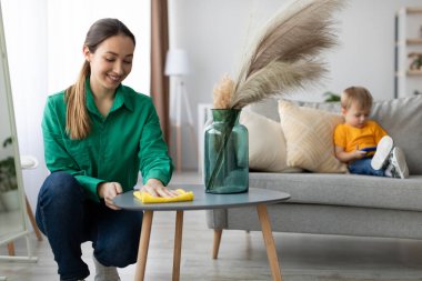 Yeşil gömlekli bir kadın sarı bir bezle kahve masasını silerken küçük çocuğu da bir cihaza odaklanmış olarak kanepede oturuyor. Oda aydınlık ve davetkar..