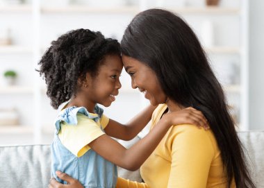 Bir anne ve kızı öne eğiliyor, alınlarına dokunuyor ve gülümsüyor. Yumuşak dekor ve ışıkla çevrili, rahat bir oturma odasında oturuyorlar. Sahne sıcaklık ve şefkat saçıyor..