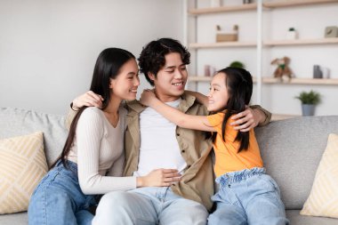 Üç aile üyesi rahat bir kanepede keyifli bir anı paylaşıyor. İki yetişkin yan yana otururken, bir çocuk onlara sevgiyle sarılıyor ve sıcak bir atmosfer yaratıyor..