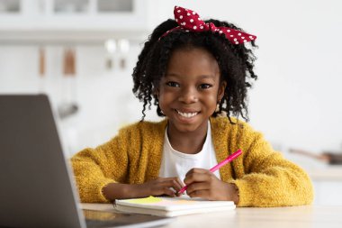 Neşeli bir kız, dizüstü bilgisayar kullanarak ve deftere yazı yazarak masa başında çalışıyor. Sarı bir kazak ve kırmızı bir benekli kafa bandı takıyor, öğrenme zamanının keyfini çıkarıyor..