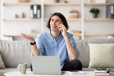 Genç bir adam kanepede oturuyor, dizüstü bilgisayarı üzerinde çalışırken gülümsüyor ve telefonla konuşuyor. Oturma odası, kitap rafları ve bitkilerle modern bir tasarıma sahip..