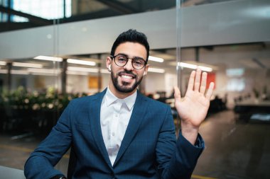 Gözlüklü ve takım elbiseli bir adam, çağdaş bir ofis yerinde masa başında otururken gülümseyip el sallıyor. Atmosfer parlak ve misafirperver. Olumlu bir hava var..