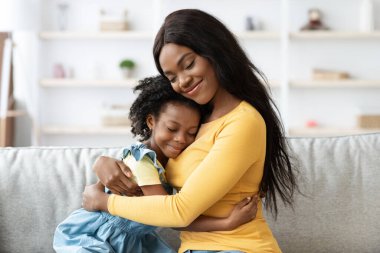 Neşeli bir anne, genç kızını rahat bir kanepeye yakın tutar. Parlak bir oturma odasında huzur içinde gülümserler. Atmosfer sevgi ve mutlulukla dolu..