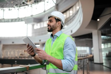 Parlak yelek ve sert şapkalı bir inşaat işçisi, büyük bir binanın içinde proje detaylarını incelerken tablet kullanarak dikiliyor..