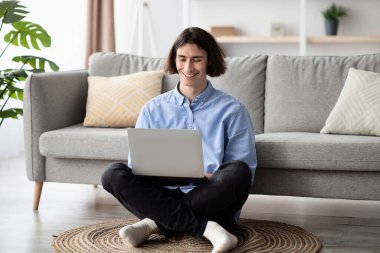 Genç bir adam şık bir oturma odasında bağdaş kurup yerde oturuyor. O, mavi gömlek ve siyah pantolon giyerken dizüstü bilgisayarına odaklanmış, gülümsüyor..