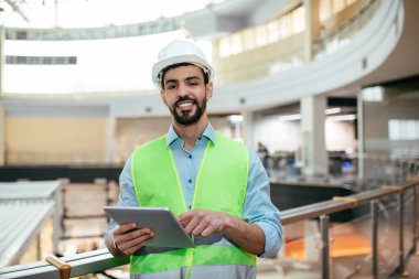 Başlık ve güvenlik yeleği giyen bir inşaat müdürü tabletle güvenle ayakta duruyor. Kapalı alanda yenilenen geniş bir tesisteki faaliyetleri izliyor..
