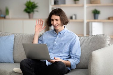 Saçı omuz hizasında genç bir adam rahat oturma odasından gelen bir video görüşmesi sırasında dizüstü bilgisayarına el sallarken gülümsüyor. Yeşil bitkiler dekoru tazeler..