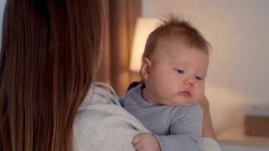 Sıcak ve davetkar bir evde, bir anne uykulu bebeğini nazikçe kucaklar. Saçları dağınık olan çocuk, kollarında gevşiyor, birlikte huzurlu bir anda sevgi bağlarını gösteriyor..