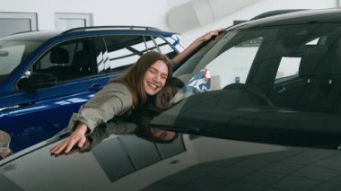 Bir araba galerisinin içindeki parlak bir arabaya yaslanırken genç bir kadın sevinçle ışıldıyor, bir araç seçme konusundaki heyecanını yansıtıyor. Atmosfer binlerce yıl boyunca canlı ve ilham verici..