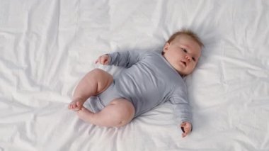 Açık gri elbiseli uykulu bir bebek, evdeki sevgi ve sıcaklıkla çevrili yumuşak beyaz bir yatakta rahatça yatar. Bu sevgi dolu an çocukluk masumiyetini ve huzurunu yakalar..