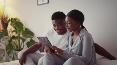 Genç bir Afrikalı Amerikalı çift rahat bir yatakta oturuyor, tabletle meşgul olurken gülümsüyorlar. Rahatlamış görünüyorlar, evde rahat bir hafta sonu boyunca birbirlerine eşlik ederek eğleniyorlar..