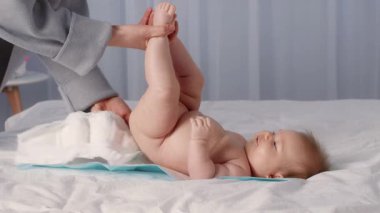Küçük bir çocuk evde bez değiştiriyor. Bakıcı bebek bacaklarını nazikçe kaldırır, sevgi dolu bir etkileşim gösterir. Samimi ortam aralarındaki bağı vurguluyor..