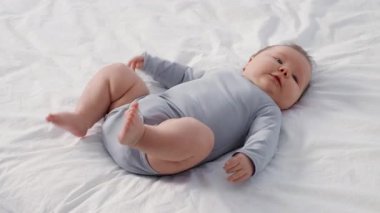 Küçük bir çocuk, evdeki sakin bir günde oynarken yumuşak bir yatak örtüsünün üzerine rahatça uzanır ve hafifçe gülümser. Sevecen atmosfer aile sevgisini ve ilgisini gösteriyor..