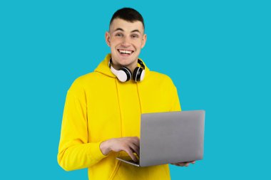 Sarı kapüşonlu ve kulaklıklı genç bir adam dizüstü bilgisayarını kullanmaktan hoşlanıyor. Neşeli ifadesi, parlak mavi bir arkaplan karşısında yaptığı faaliyetleri yansıtıyor..