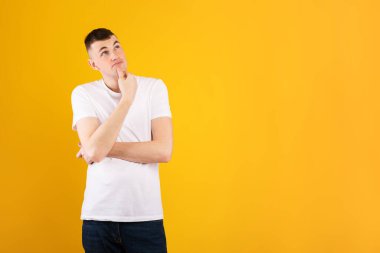 Beyaz gömlekli genç bir adam kollarını kavuşturmuş, düşünceli bir şekilde yukarı bakıyor. Parlak sarı arka plan, sahneye canlı ve neşeli bir atmosfer ekler..