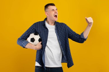 Neşeli genç bir adam futbol zaferinden sonra heyecanını ifade eder. Bir elinde siyah beyaz bir balo tutuyor ve kutlama için yumruğunu kaldırıyor. Parlak sarı arkaplan mutlu anı güçlendirir.