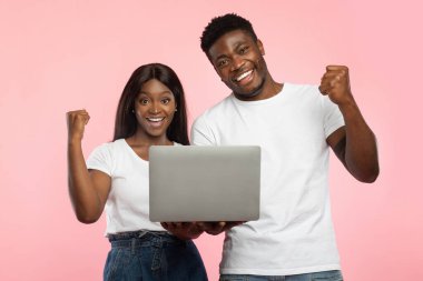 Heyecanlı bir siyah çift pembe bir duvarın önünde duruyor, online iş başarılarını kutluyorlar. Sevinçle el kol hareketi yapıyorlar, yumruklarını havaya sallıyorlar ve ellerinde bir dizüstü bilgisayar tutuyorlar..