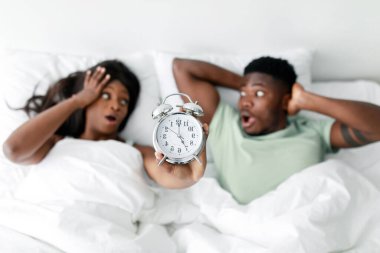 A couple is in bed with a shocked expression as one holds an alarm clock. They appear to have just woken up and are reacting to the late hour.