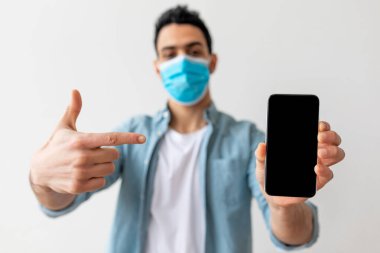 Mavi maske takan genç bir adam bir elinde akıllı bir telefon tutarken diğer elinde onu işaret ediyor. Ortam modern ve aydınlık görünüyor. Bu da sıradan bir atmosfer olduğunu gösteriyor..