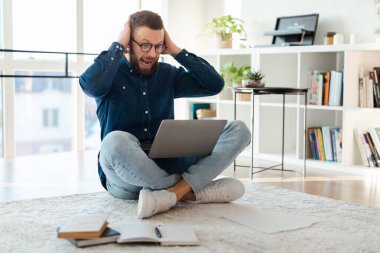 Yerde bacakları çapraz oturan bir adam, iyi aydınlatılmış bir oturma odasında dizüstü bilgisayar kullanırken şaşırmış ve heyecanlı görünüyor. Kitaplar ve kağıtlar yakınlarda..