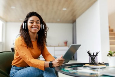 Genç bir kadın bir sandalyede rahatça otururken, kulaklık takarak ve tablet tutarak gülümsüyor. Parlak bir oturma odasında şık bir dekorasyonu ve küçük bir masası var..