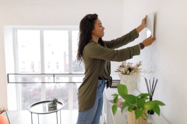 Genç bir kadın, parlak ve modern bir yaşam alanındaki açık renkli bir duvara bir sanat eseri asıyor. Dekoru hazırlarken gülümsüyor..