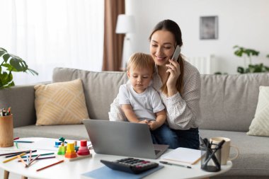 A woman is engaged in a phone call while seated on a couch. Her young child is sitting on her lap, playing with a laptop. Colorful toys and stationery are on the table nearby.
