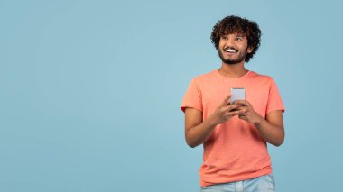 Neşeli bir Hintli adam cep telefonunu kullanırken kameraya bakar. Meşgul görünüyor, muhtemelen arkadaşlarıyla sohbet ediyor ya da oyun oynuyor..