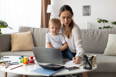Bir anne, küçük oğlu yanında otururken bir dizüstü bilgisayarla masaya oturur ve notlar yazar. Oturma odasında bitkiler ve sanat yer alıyor, çalışmak ve oynamak için sıcak bir atmosfer yaratıyor..