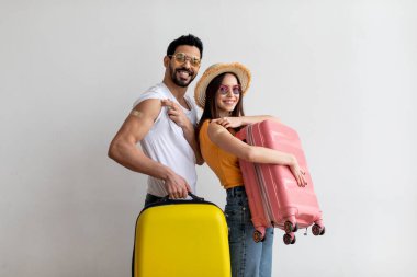 Neşeli bir çift, güneş gözlüğü ve yaz kıyafetleri takan iki bavulla poz veriyor. Erkeklerin kolunda yara bandı görülüyor. Bu da yeni aşıyı ya da korunmayı akla getiriyor..