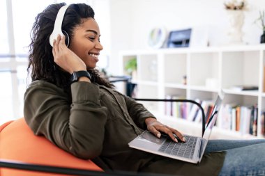 Bir kadın dizüstü bilgisayarı açık, kulaklık takmış ve gülümsüyor. O, kitaplarla dolu parlak, davetkar bir oturma odasında işi veya boş zamanı ile meşgul..