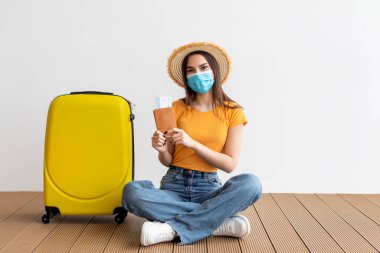 Genç bir kadın seyahat belgelerini tutarken dizlerinin üstüne çöküp gülümsüyor. Yanında sarı bir bavul ve hasır şapka takıyor..