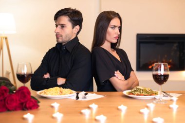 Bir çift yemek masasında sırt sırta oturuyor. Görünürde üzgün ve göz temasından kaçınıyor. Etrafları romantik dekorlarla çevrili ama anlaşmazlıkları ortamı gölgeliyor..