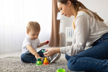 Genç bir çocuk ve bir anne renkli oyuncakları birlikte istiflemekten zevk alırken yumuşak bir halıya otururlar. Parlak odada doğal ışık var. Öğrenmek ve oynamak için sıcak bir atmosfer yaratıyor..