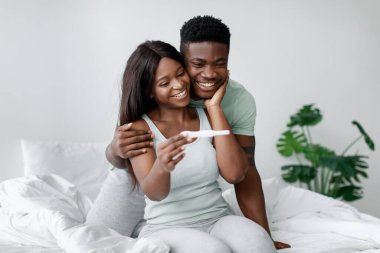 Mutlu bir çift, pozitif bir hamilelik testi yaparken yataklarında oturup gülümserler. Parlak oda modern dekor ve yeşilliğe sahip, sıcak bir atmosfer yaratıyor..