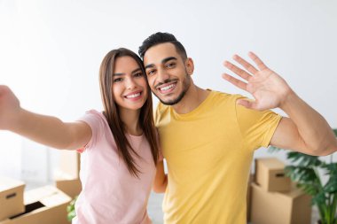 Neşeli bir çift yeni yaşam alanlarında taşınan kutularla dolu bir selfie için poz veriyor. Gülümseyip el sallıyorlar, yeni başlangıçlarını kutluyorlar..
