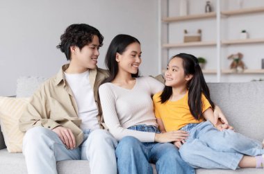 Genç bir aile, parlak bir oturma odasındaki kanepede birlikte oturur. Gülümseyen ebeveynler ve kızları günlük kıyafetlerini giyerken neşeli bir anı paylaşıyorlar..