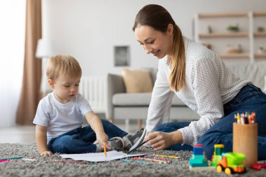 Mother and son are sitting on a soft carpet in a cozy living room. The boy is excitedly pointing at colorful drawings while the mother encourages him.
