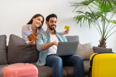A joyful couple sits on a couch, excitedly planning their next vacation. They use a laptop while holding travel documents and credit cards, surrounded by colorful luggage.