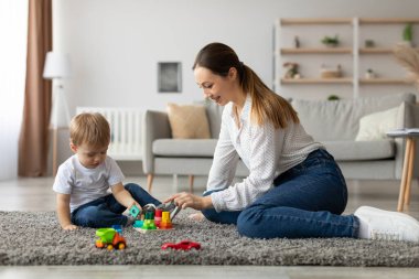 Genç bir çocuk yumuşak bir halıya oturur, renkli bloklarla inşa etmeye odaklanır. Bu sırada annesi ona yardımcı olur, keyifli oturma odalarında besleyici bir ortam yaratır..